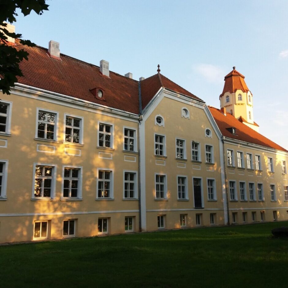 Plašs gaiši dzeltenas krāsas nams ar sarkanu dakstiņu jumtu un torni. Nams atrodas zaļā zālienā, un saules gaisma rada ēnas uz tā fasādes. Tālumā redzama sarkanīgi oranža ēka.