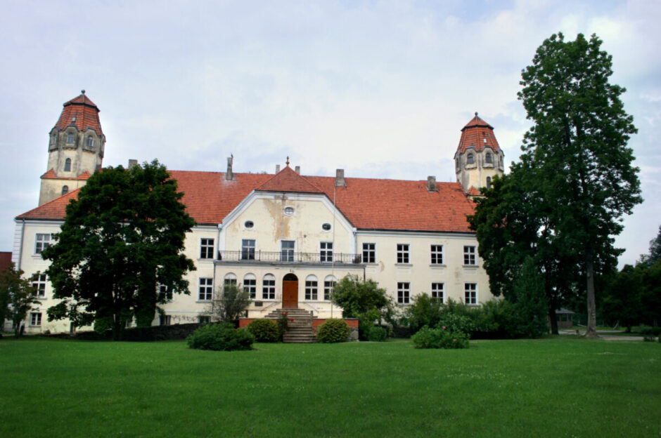 Gaišas krāsas vēsturiska ēka ar sarkanu dakstiņu jumtu un diviem torņiem ar konusveida jumtiem sānos. Priekšplānā plašs zaļš zāliens un lieli koki.